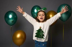 schön wenig Mädchen tragen ein Weiß Weihnachten Sweatshirt und Elf Band, erhöht ihr Waffen oben Stehen gegen bunt golden und Grün schön Luft Luftballons isoliert Über grau Hintergrund mit Kopieren Raum foto