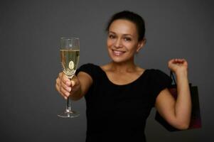 Fokus auf Champagner Flöte, Glas mit funkelnd Wein im das Hände von ein lächelnd schön jung Frau gekleidet im schwarz und halten ein schwarz Einkaufen Tasche, feiern schwarz Freitag. Kopieren Raum zum Anzeige foto