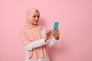 isoliert Porträt von zuversichtlich arabisch Muslim Mitte alt Frau im streng religiös Outfit und bedeckt Kopf im Rosa Hijab SMS ein Botschaft auf ein Handy, Mobiltelefon Telefon im ihr Hände, farbig Hintergrund, Kopieren Raum foto