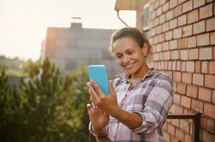 heiter Frau mit Smartphone, Herstellung Selfie Stehen auf das Balkon von ein Land Seite Haus auf sonnig warm Sommer- Tag foto