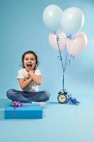 glücklich wenig Mädchen ausdrücken ein positiv Emotion, Glück und Befriedigung Sitzung auf Blau Hintergrund in der Nähe von ein Alarm Uhr und Farbe Luftballons und ein Blau Geschenk im ein Vorderseite von ihr foto