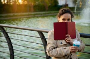 heiter Frau Abdeckungen ihr Gesicht mit ein gebundene Ausgabe Buch, sieht aus zu das Seite, Stehen gegen das Hintergrund von ein See, halten ein Tasse mit heiß Kaffee. Herbst Hintergrund, draussen Aktivitäten, sich ausruhen von Gadgets foto