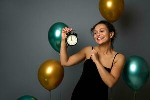 lächelnd Frau im schwarz Kleid, freut sich beim Party, sieht aus beim das Alarm Uhr im ihr Hand, es ist Mitternacht. feiern Weihnachten, Neu Jahr, Geburtstag Konzept auf grau Hintergrund mit Kopieren Raum foto