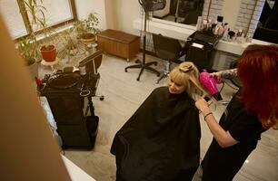 hoch Winkel Aussicht von Rothaarige weiblich Friseur Kämmen und Trocknen Klient Haar im Schönheit Salon, mit Fön zu ausführen stilvoll Haar Stil. Mode, klein Geschäft und Schönheit Industrie Konzept foto