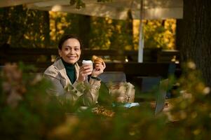 attraktiv dunkelhaarig Geschäft Frau tragen Beige Mantel haben ein Kaffee brechen im ein draussen Terrasse von ein hölzern Cafeteria im Eiche Hain auf schön sonnig Herbst Tag. beschäftigt Frau ruhen nach Arbeit foto