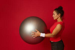 Seite Porträt von ein schön Sportlerin ausüben mit Fitness Ball während tun ein funktional Kreuz und Körper Gewicht Ausbildung zum Abnehmen und Gewicht Verlust, gegen rot Hintergrund mit Kopieren Anzeige Raum foto