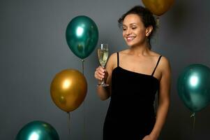 fröhlich Weihnachten und glücklich Neu Jahr. Werbung Schuss von ein schön spanisch Frau im Abend Kleid mit Glas von Champagner posieren gegen grau Hintergrund mit glänzend Gold und Grün metallisch Luftballons foto