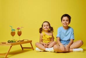 zwei bezaubernd Kinder Sitzung im Lotus Position in der Nähe von ein hölzern Portion Tablett mit Cocktails und Teller mit geschnitten Früchte foto