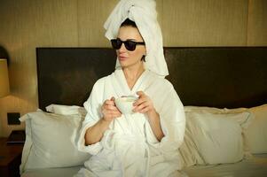 attraktiv Frau im Sonnenbrille und ein Weiß Frottee Bademantel mit ihr Kopf eingewickelt im ein Weiß Bad Handtuch, halten ein Tasse von Kaffee und entspannend im das Schlafzimmer, Sitzung auf das Bett und suchen aisde foto