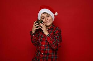 schön zärtlich süß wenig Junge, bezaubernd Kind im Santa Hut und kariert Hemd sanft Umarmungen seine Weihnachten Geschenk im Grün Verpackung Papier mit golden Bogen, isoliert auf rot Hintergrund mit Kopieren Raum foto