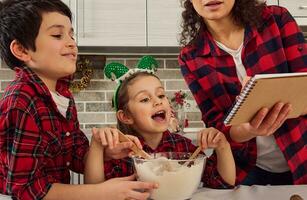 schön wenig Mädchen mit Elf Band freut sich, Sitzung beim ein Tabelle zwischen ihr gut aussehend vorpubertär Bruder und schön Mutter während Kochen zusammen ein Weihnachten Kuchen beim Zuhause Küche foto