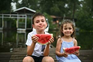 bezaubernd Kinder, Kinder, Junge und Mädchen Sitzung schließen zu jeder andere mit Scheibe von köstlich saftig Wassermelone im Hände und süß lächelnd zu Kamera auf das Fluss Hintergrund foto