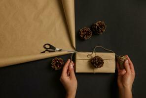Mensch Hände halten einfach rustikal Geschenk im Kunst Verpackung Papier dekoriert mit ein Kiefer Kegel und gebunden Bogen von ein Seil. Frau dekorieren Weihnachten die Geschenke. Weihnachten Vorbereitung . foto