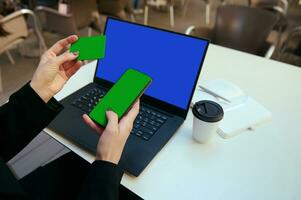 Nahansicht. Hände halten Grün leer leeren Anerkennung Rabatt Karte und Smartphone mit Chroma Schlüssel zum Platzierung Anzeigen und Einkaufen online im Vorderseite von Laptop mit Blau Attrappe, Lehrmodell, Simulation Monitor Bildschirm. online Geschäft foto