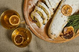 eben legen. frisch gebacken traditionell hausgemacht Deutsche Stollen Brot bestreut mit Glasur Zucker, aufwendig mit Seil, Orange Scheiben, Kiefer Zweige auf ein hölzern Planke, Tassen mit gesund Kräuter- trinken mit Nelken foto