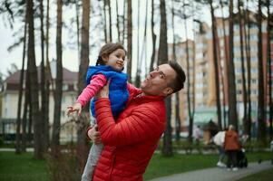 gut aussehend kaukasisch Mann - - liebend Papa wirft seine Tochter - - ein wenig süß Mädchen hoch oben im ein Stadt Park. Familie und Vaters Tag, Liebe, Eltern, Kinder foto