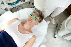 ein Overhead Aussicht von ein Klient von ein Kosmetologie Klinik im schützend ultraviolett Brille Lügen auf ein Massage Tabelle Vor ein Epilation Verfahren mit ein modern Laser- Maschine foto