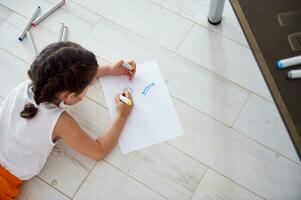 Aussicht von über Vorschulkind Mädchen Zeichnung Bild auf Weiß Papier Blatt mit Aquarell Filzstifte, Lügen auf das Fußboden foto