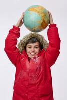 lächelnd heiter Junge im hell rot schneebedeckt Nieder Jacke mit Haube, halten enorm Globus im seine Hände über seine Kopf, isoliert auf Weiß Hintergrund. Tourismus, Erdkunde, Planet Erkundung Konzept. Kopieren Raum foto