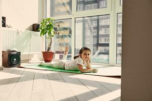schön wenig Mädchen ruhen auf ein Yoga Matte gegen das Hintergrund von groß Fenster beim Zuhause foto