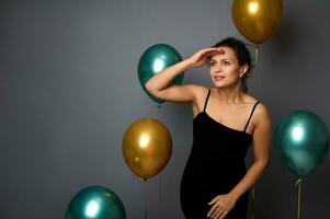 charmant Mitte alt Frau im Abend elegant Kleid hält ihr Hand auf ihr Tempel, sieht aus weg, in Entfernung mit nachdenklich sehen. isoliert Über grau Hintergrund mit golden Grün Luftballons auf Kopieren Raum foto