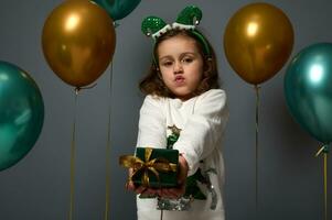 bezaubernd wenig Mädchen im festlich Sweatshirt mit glänzend Weihnachten Baum und Elf Band auf ihr Kopf posiert auf grau Hintergrund mit goldgrün glänzend Luft Luftballons und zeigt an ein Luxus klein Geschenk Box zu das Kamera foto