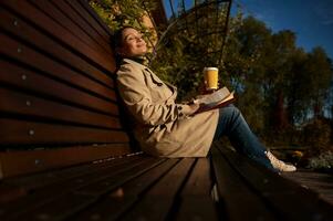 attraktiv Frau mit geschlossen Augen sitzt auf hölzern Bank im Park, halten Papier Tasse von heiß trinken und ein Buch, genießt Herbst Sonnenstrahlen, Weg von das Gedränge und Geschäftigkeit von das Stadt, ruhen von Gadgets foto