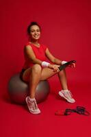 charmant atemberaubend spanisch Sportlerin halten elastisch Fitness Band und Sitzung auf Fitness Ball nach ausüben isoliert auf rot Hintergrund. Konzept von schlankheit, gesund Lebensstil und trainieren. foto