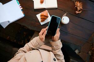 Nahansicht von ein unkenntlich Person Hände mit ein Handy, Mobiltelefon Telefon mit leer schwarz Bildschirm zum Kopieren Raum auf das Hintergrund von ein hölzern Tabelle mit öffnen Notizblock und Heft und köstlich Frühstück foto