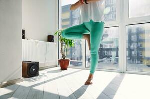 Nahansicht von jung schön sportlich Frau üben Yoga und durchführen Baum Pose beim Zuhause gegen groß Fenster foto