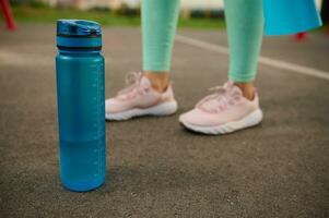 Nahansicht von ein Blau Plastik Flasche von Wasser auf das Asphalt von Sport Boden Nächster zu sportlich Frau Füße. Gesundheit und Körper Pflege Konzept foto