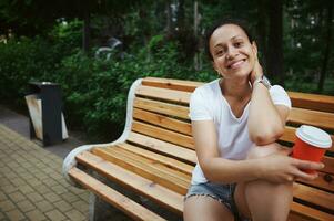 schön Frau lächelnd beim Kamera, halten ein Karton Becher mit wegbringen Kaffee, Sitzung auf hölzern Bank im das Park foto