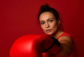 Nahansicht Porträt von konzentriert jung Sport Frau Boxer tragen rot Boxen Handschuhe, Herstellung Direkte Schlag, Stanzen gegenüber Kamera, isoliert Über rot Hintergrund mit Kopieren Raum. kriegerisch Kunst Konzept foto