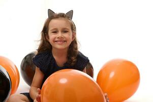 Porträt von süß Hexe Mädchen im Halloween Kleid und Band mit Katze Ohren haben Spaß mit bunt schwarz und Orange Luftballons isoliert auf Weiß Hintergrund mit Kopieren Raum. Halloween Party. foto