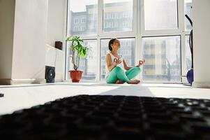 jung Frau meditieren im das Lotus Position gegen das Hintergrund von groß Fenster im das Zuhause Fitnessstudio. foto