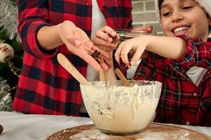 Nahansicht von Mamas und ihr Söhne Hände Gießen etwas Rosinen in ein Schüssel mit Teig während Kochen zusammen im das Zuhause Küche foto