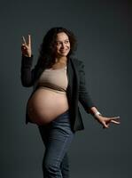 heiter lächelnd Latein amerikanisch schwanger Frau zeigt an Frieden Zeichen, posiert nackt Bauch Über grau isoliert Studio Hintergrund foto