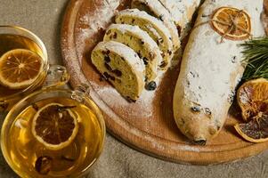 oben Aussicht von frisch gebacken traditionell Deutsche Stollen Brot bestreut mit Glasur Zucker, aufwendig mit ein Seil und getrocknet Orange Scheibe auf ein hölzern Tafel in der Nähe von ein Glas Tasse mit heiß Kräuter- Tee, Kiefer Zweige foto