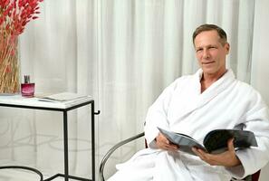 gut aussehend reifen kaukasisch Mann gekleidet im Weiß Frottee Bademantel, selbstbewusst suchen beim Kamera während entspannend im Privat Zimmer von ein Wellness Spa Center, Sitzung auf ein Sessel mit Zeitschrift im seine Hände foto