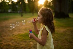 süß Mädchen 4 Jahre alt, weht Seife Luftblasen gegen ein Stadt Park Hintergrund beim Sonnenuntergang, genießen angenehm Zeit draußen. Sonne Strahlen fallen durch transparent Blase Kugeln mit irisierend Reflexionen. foto