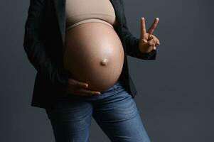 Mittelteil schwanger Frau halten ihr groß Bauch, zeigen zwei Finger, erwarten Zwillinge oder gestikulieren mit ein Frieden Zeichen foto