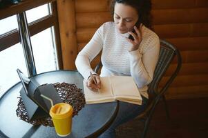 jung lockig Frau konzentriert beim arbeiten, Schreiben auf Tagebuch, reden durch Smartphone während Sitzung in der Nähe von Fenster im ein hölzern Cafeteria foto