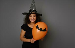 heiter jung Frau im schwarz und Magier Hut posiert mit ein Orange Luft Ballon mit ein handgemacht Filzschnitt Schläger, lächelt zahnig Lächeln suchen beim Kamera. Halloween Konzept auf grau Hintergrund, Kopieren Raum foto