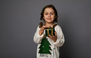 schön wenig Mädchen im Weiß Sweatshirt mit ein Weihnachten Baum, posiert gegen grau Hintergrund, halten ein Weihnachten Geschenk im Grün funkeln Verpackung Papier und golden Band, süß lächelt suchen beim Kamera foto