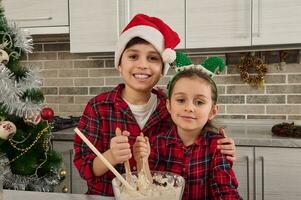gut aussehend kaukasisch Junge im Santa claus Hut lächelt mit heiter zahnig Lächeln umarmen seine bezaubernd wenig Schwester und suchen beim Kamera während kneten und vorbereiten Teig zum Weihnachten traditionell Kuchen foto