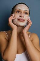 charmant Frau bewirbt sich Schaum Reinigung kosmetisch Produkt auf ihr Gesicht, tun Massage Bewegungen, entfernen bilden und erfrischend ihr Haut mit ein Peeling Schönheit Produkt, isoliert Über grau Hintergrund foto