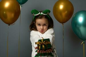 süß Mädchen im gemütlich Weiß Sweatshirt mit glänzend Weihnachten Baum und Elf Band auf ihr Kopf posiert auf grau Hintergrund dekoriert mit goldgrün glänzend Luftballons und zeigt an ein Luxus klein Geschenk Box zu das Kamera foto