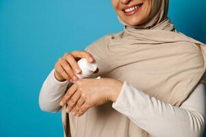 Nahansicht von Frau im Hijab quetschen Sahne auf Hand, isoliert auf Blau Hintergrund mit Raum zum Text foto