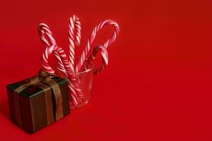 Weihnachten Dekoration zum Anzeige. glänzend Geschenk im funkeln Grün Verpackung Papier mit golden Bogen in der Nähe von Glas mit gestreift Lutscher, isoliert Über rot Hintergrund mit Kopieren Raum foto