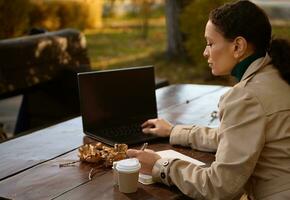 Porträt von ein ziemlich Frau, zuversichtlich Geschäft Dame Arbeiten auf Laptop und Schreiben auf das wöchentlich während nehmen Frühstück im draussen Wald Cafe im das Eiche Hain auf warm Herbst Tag foto